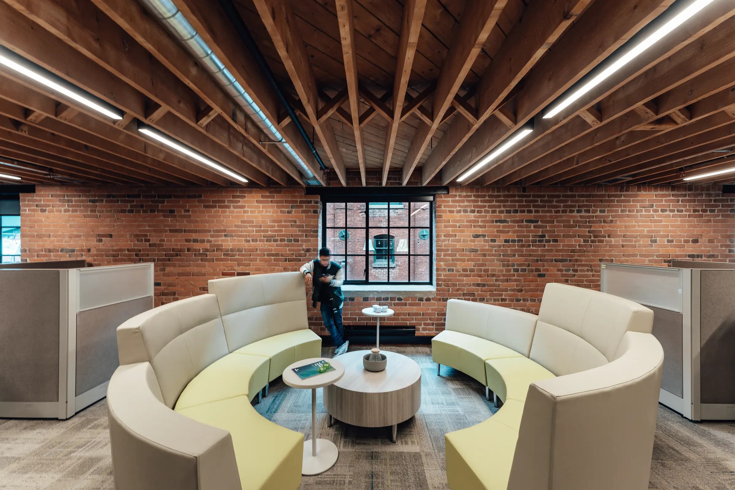 A student lounge at Collège Boréal under exposed timber joists and a red-brick wall: two curved banquettes in cream and lime face each other around a round wood-grain table, framed by workstation panels, with a student leaning by a multi-paned window.
